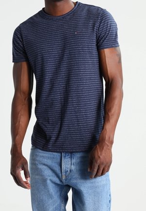 Mann trägt ein eng anliegendes marineblau gestreiftes T-Shirt und helle blaue Jeans, steht vor einem schlichten weißen Hintergrund.