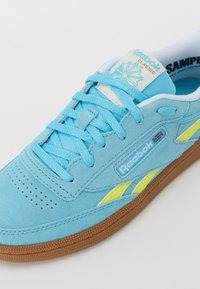 Sapatilhas de camurça azul claro com detalhes amarelos, atacadores planos, forro têxtil, bico perfurado e sola de borracha castanha. Logotipo da Reebok em destaque.