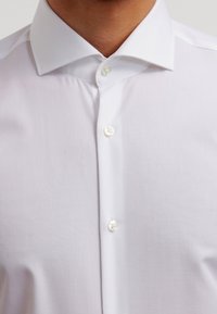 White dress shirt with a pointed collar, made of smooth fabric; features pearl buttons along the front for closure. Simple and clean design.