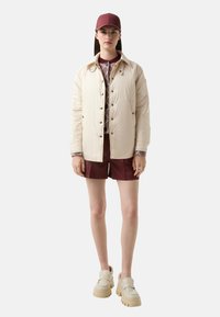 Jasno beżowa kurtka puchowa z zapięciem na guziki, noszona na wzorzystej bluzce i bordowych szortach. W komplecie z kremowymi butami na grubej podeszwie i bordową czapką.