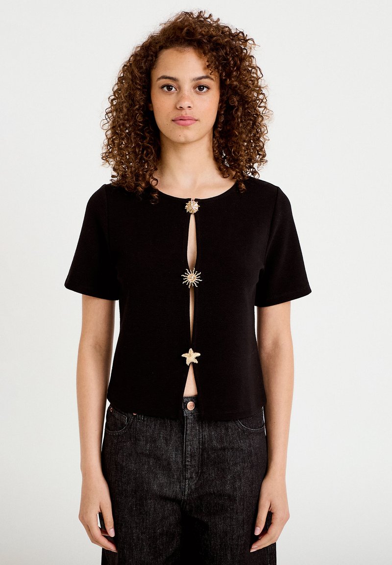 Mujer con cabello rizado que lleva una camiseta negra de manga corta con tres broches decorativos en forma de sol y estrella en la parte delantera y vaqueros negros.