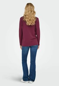 Kastanis sarkans garām piedurknēm tops ar taisnu griezumu, apvienots ar platām denima biksēm. Aizmugurējais skats rāda gludu audumu un dabiski viļņotus matus.