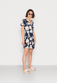 Vestido curto florido em azul marinho com flores em rosa claro e branco, decote em V, mangas bufantes. Combinado com sandálias de plataforma bege, com palmilha. 