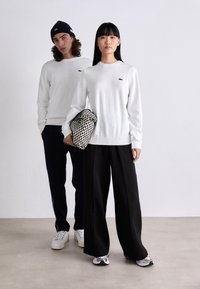 White knitted sweater with a small logo, paired with black wide-leg trousers. The woman holds a patterned, net-style bag. The man wears navy pants.