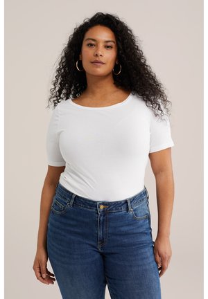 Femeie cu păr creț, purtând cercei aurii rotunzi, un top alb cu mâneci scurte și blugi albaștri cu talie înaltă, stând în picioare pe un fundal simplu.