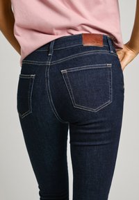 Slim fit, sötét farmernadrág magas derékkal, kontrasztos varrással és két hátsó zsebből áll. Bőr márkavédjegy a derékpánton.
