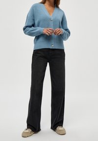 Cardigan lavorato a maglia di un blu chiaro, con scollatura a V e maniche oversize, dotato di un singolo bottone decorativo e indossato sopra pantaloni neri a gamba larga.