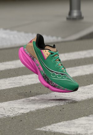 Πράσινο και μπεζ αθλητικό παπούτσι με ροζ σόλα στον αέρα πάνω από διάβαση πεζών σε έναν δρόμο με θολό φόντο.