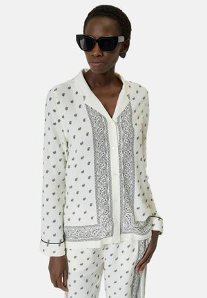 Kobieta w białym stroju w stylu piżamy z czarnym wzorem paisley, dużymi czarnymi okularami przeciwsłonecznymi i czarnymi kolczykami w kształcie serca.