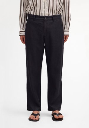 Persona che indossa pantaloni neri a gamba dritta, camicia a righe con bottoni infilata dentro e sandali infradito neri, in piedi su sfondo bianco.