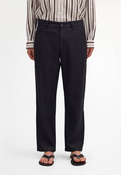 Osoba stojící na bílém pozadí, oblečená v černých kalhotách s rovnými nohavicemi, pruhované košili s knoflíky zastrčené dovnitř a černých žabkách.