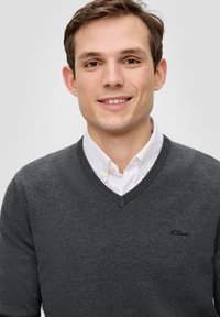 Grauer V-Ausschnitt-Pullover mit geripptem Kragen, getragen über einem weißen Hemd mit Kragen. Verfügt über ein dezentes Logo auf der linken Brust und eine glatte Textur.