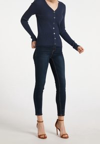 Granatowy sweter z prążkowanej dzianiny z dekoltem w serek i zapięciem na guziki, zestawiony z ciemnoniebieskimi dżinsami typu skinny oraz brązowymi sandałami na obcasie.