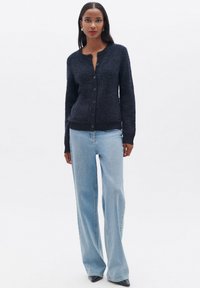 Cardigan navy a trama con chiusura a bottoni, collo rotondo e polsini a coste, abbinato a jeans a gamba larga blu chiaro e scarpe nere a punta.