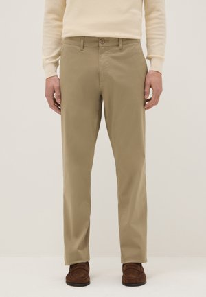 Uomo con pantaloni beige a gamba dritta, maglione crema a maniche lunghe e mocassini marroni in suede, in piedi su uno sfondo neutro.