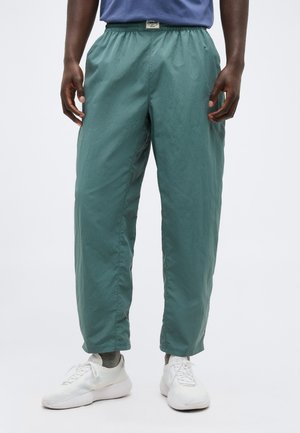 Άνδρας που φοράει καφέ χαλαρό παντελόνι με ελαστική μέση, γκρι κάλτσες και λευκά αθλητικά παπούτσια σε ανοιχτό φόντο.