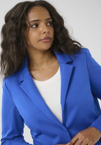 Blauwe blazer met een gelapte revers, gladde stof, enkele knoopsluiting en een getailleerde pasvorm, gedragen over een witte top.