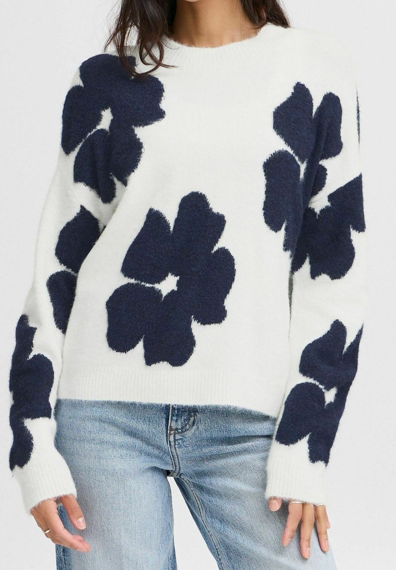 Femme portant un pull blanc duveteux avec de grands motifs floraux bleu marine et un jean bleu clair, vue du cou aux hanches.