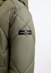 Olivengroene gewatteerde pufferjack met een mouw die voorzien is van een zwart logo-embleem met de tekst "FRIEDA & FREDDIES NEW YORK." Soepele textuur, lichtgewicht ontwerp.