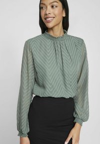 Jasnozielona bluzka z długimi rękawami, marszczonym dekoltem, wzorem w zygzak oraz elastycznymi mankietami. Połączona z czarną spódnicą.