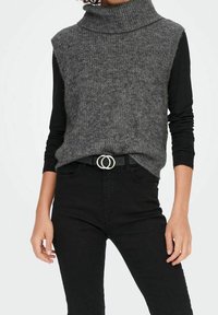 Personne portant un pull en laine gris sans manches avec col roulé par-dessus un haut à manches longues noir, un pantalon noir et une ceinture noire avec une boucle double.