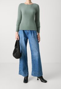 Femme portant un pull vert côtelé, un jean bleu à jambes larges, des bottines noires à talons, et tenant un sac à main noir tressé.