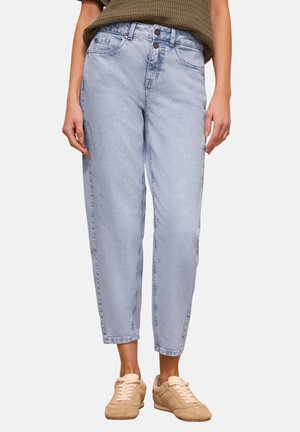 Personne portant un jean en denim bleu clair taille haute et des baskets beige, debout avec les mains détendues le long du corps devant un fond uni.