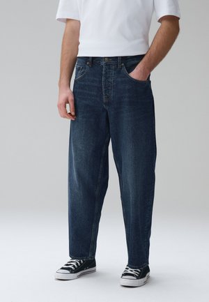 Mand iført løstsiddende mørkeblå jeans, hvid T-shirt med korte ærmer, sorte og hvide sneakers, med den ene hånd i lommen mod en ensfarvet baggrund.