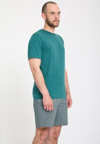 Homme barbu portant une chemise turquoise à manches courtes et un short gris, debout de profil sur un fond blanc uni.