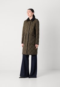 Olijfgroene gewatteerde lange jas met een capuchon, zwarte kraag, strikceintuur in de taille en twee zakken aan de voorkant; gecombineerd met een wijdvallende donkerdenim broek.