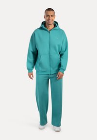 Turkusowa bluza z zamkiem, z dużą przednią kieszenią, w komplecie z dopasowanymi spodniami dresowymi. Charakteryzuje się luźnym krojem i ściągaczami na końcach rękawów.