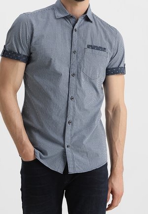 Homme portant une chemise à manches courtes boutonnée avec un motif à carreaux bleu marine et blanc et des manches retroussées avec un tissu bleu marine contrastant.