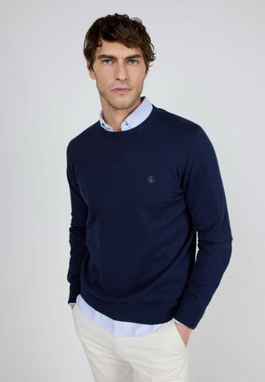 Joven con cabello castaño ondulado que lleva un jersey azul marino sobre una camisa de cuello azul claro y pantalones blancos con las manos en los bolsillos.