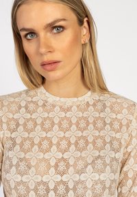 Vrouw met lang blond haar draagt een crèmekleurige kanten top met bloemenpatroon en toont meerdere kleine oorbellen in haar rechteroor.