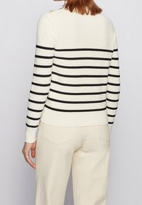 Pull couleur crème avec des rayures horizontales noires, texture côtelée et détails de boutons aux épaules. Porté avec un pantalon de couleur claire.