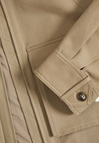 Beige jas met een gewatteerde voering, voorzien van een opstaande kraag, knoopsluiting bij de manchetten en een patch pocket aan de linkerkant.