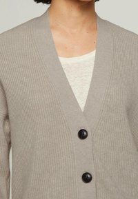 Cárdigan beige confeccionado con tela de punto texturizada, con diseño de escote en V y dos grandes botones negros al frente. Debajo lleva una camiseta de color claro.