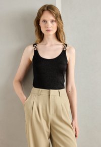 Black glitter tank top with adjustable straps featuring round hardware details, paired with beige high-waisted trousers. Smooth fabric texture.