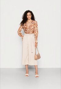 Blusa de padrão floral com mangas longas, combinada com calças largas bege de cintura alta. Acentuada com uma pequena mala e sandálias de deslizar.