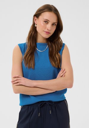 Jeune femme aux longs cheveux bruns portant un haut bleu sans manches, collier et boucles d'oreilles en argent, bras croisés, fond neutre.