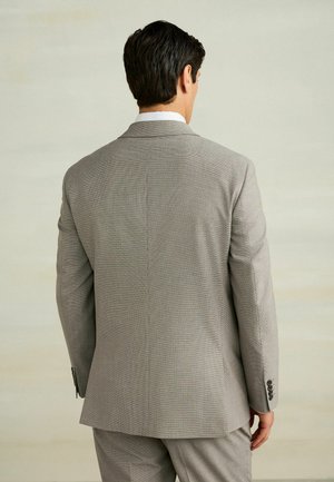 Mężczyzna o ciemnych włosach stoi tyłem, ubrany w jasnoszary, teksturowany garnitur i pasujące spodnie na neutralnym tle.