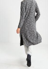 Cárdigan largo de punto gris-negro con un patrón texturizado, aberturas laterales y corte recto, llevado sobre pantalones de colores claros y botas de tobillo.