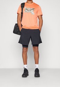 Orange Nike T-shirt med grafisk design, kombineret med sorte træningsshorts. Tilbehør inkluderer en sort taske og sorte sneakers. Hvide sokker er synlige.