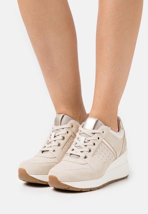 Beige wedgesneakers met veters, gedragen aan de voeten, met geperforeerd suède en metalen accenten op een witte achtergrond.