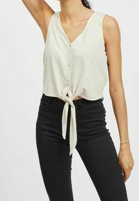 Light beige sleeveless top with a V-neck and front tie detail, made of soft fabric, paired with fitted black jeans.