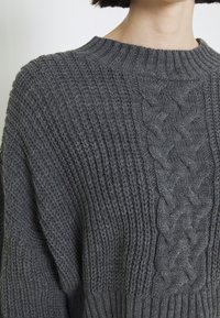 Pull en maille gris avec une texture côtelée et un motif en torsades sur le devant. Il a un col rond et une coupe décontractée.