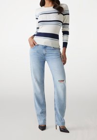 Suéter de punto en color crema con rayas en azul marino y azul, con un logo de "GUESS" adornado con lentejuelas. Jeans desgastados en azul claro y zapatos negros de tacón.