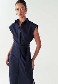 Abito camicia blu navy con maniche corte a cappuccio, chiusura a bottoni davanti, cintura in vita e spacchi laterali. Tessuto morbido con una silhouette strutturata.