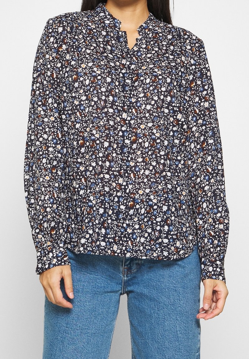 Blouse fleurie avec un fond bleu marine, ornée de fleurs blanches, bleues et rouille ; dotée d'un col montant et de manches longues.