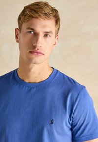 Niebieska bawełniana koszulka z okrągłym dekoltem i krótkimi rękawami. Posiada małe haftowane logo na lewej piersi. Gładka tekstura.